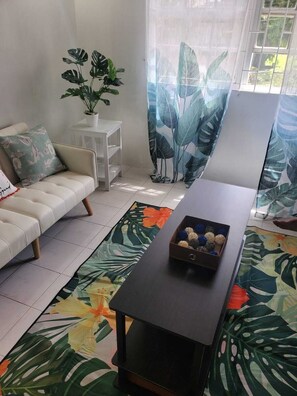 Uma sala de estar com um sofá, uma pequena mesa branca, uma mesa de centro de madeira escura com objetos decorativos e uma janela grande com cortinas.