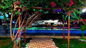 Un espacio al aire libre de noche con un sendero pavimentado, una piscina y un edificio al fondo.