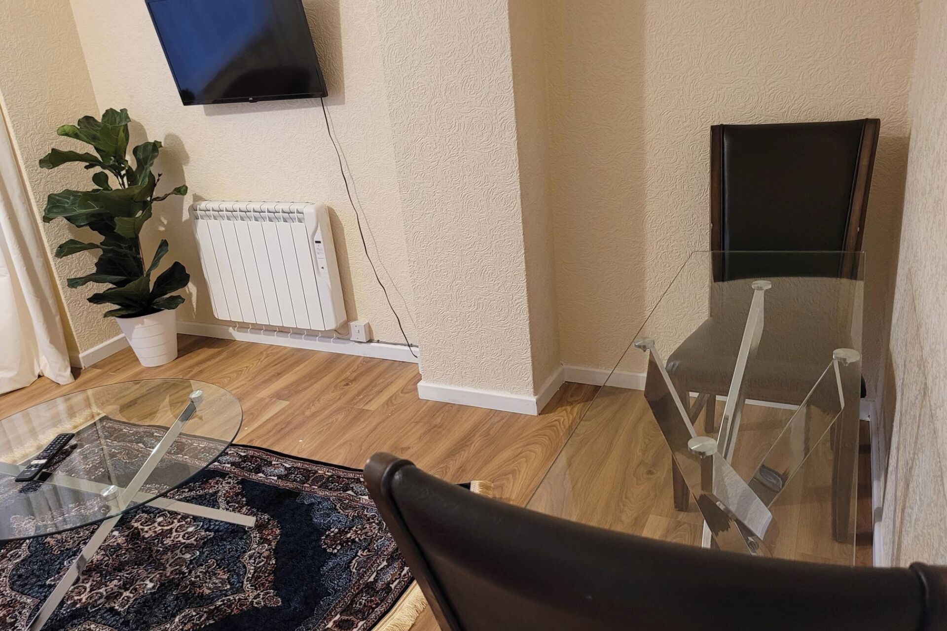 Quarto com radiador, planta em vaso, mesa de vidro e tapete estampado, além de uma cadeira.