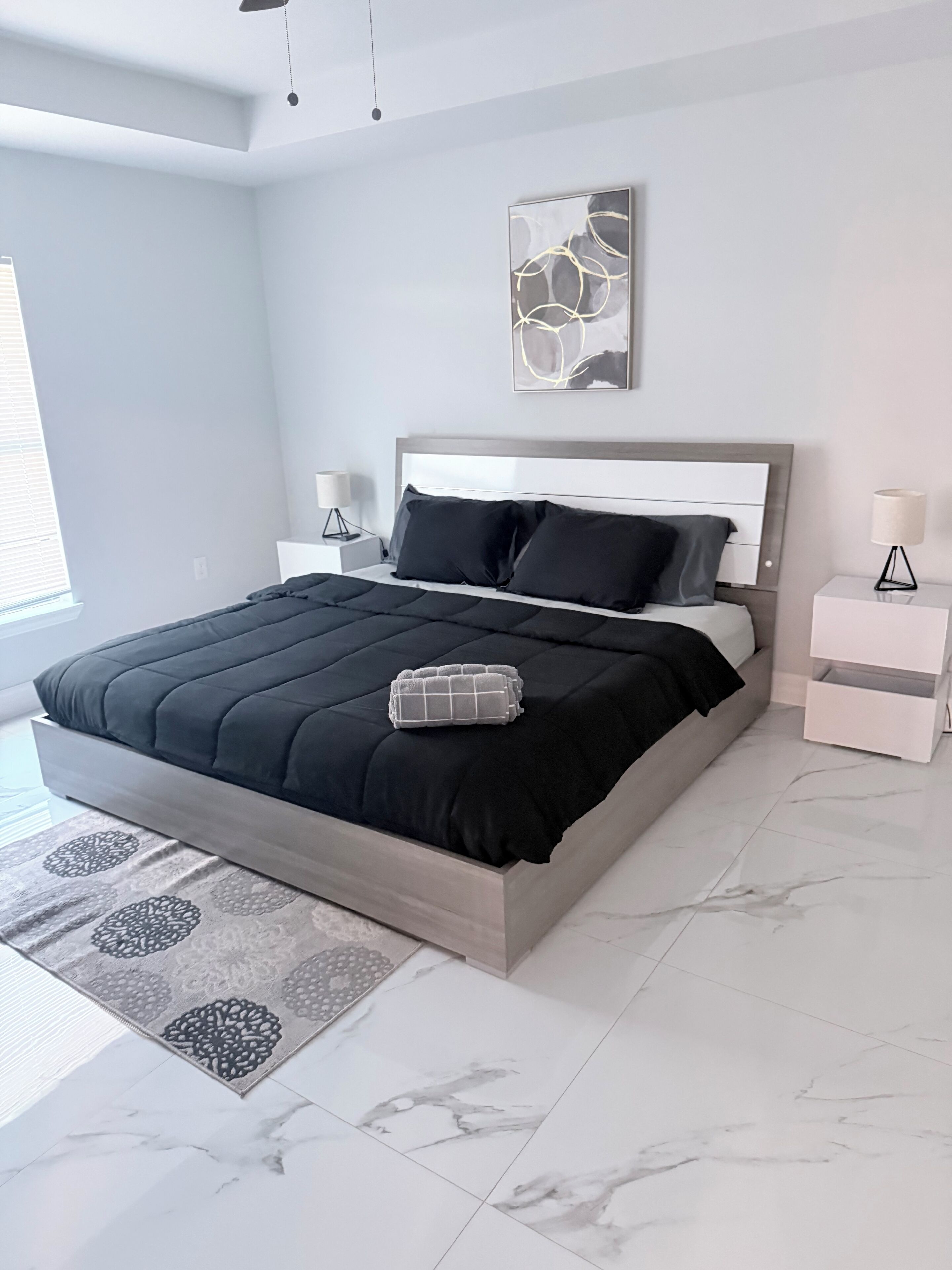 A modern bedroom with a large bed, a headboard, two bedside tables, a floor lamp, and a wall art piece.