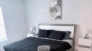 A modern bedroom with a large bed, a headboard, two bedside tables, a floor lamp, and a wall art piece. - Newly built home near SWIFlorida airport  (Lehigh Acres)