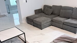 A modern living room with a grey sectional sofa, a glass-top coffee table, and a marble-patterned rug. - Newly built home near SWIFlorida airport  (Lehigh Acres)
