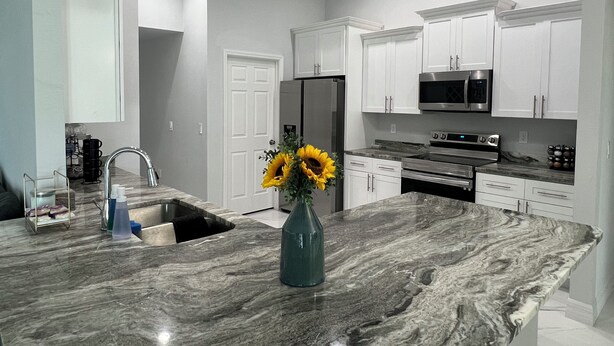 A modern kitchen with white cabinetry, a marble island, stainless steel appliances, and a vase with sunflowers. - Newly built home near SWIFlorida airport  (Lehigh Acres)
