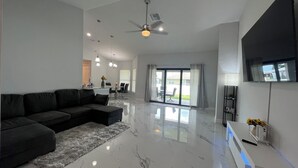 A spacious living area with a large sectional sofa, a flat-screen TV mounted on the wall, and a ceiling fan. - Newly built home near SWIFlorida airport  (Lehigh Acres)