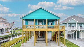 Une maison turquoise à deux étages, dotée d’un balcon, d’une terrasse en bois et d’un petit quai s’avançant dans une étendue d’eau.