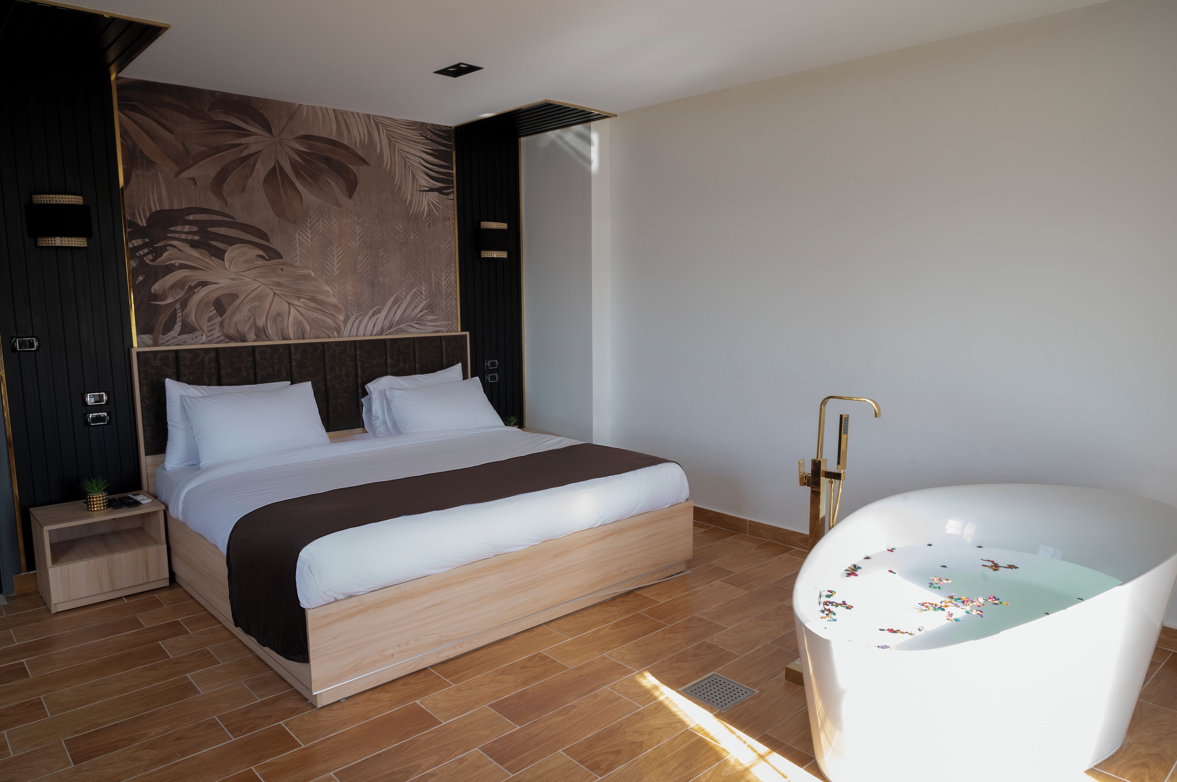 A modern bedroom with a large bed, a built-in nightstand, a wall-mounted headboard, and a freestanding bathtub.
