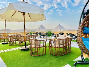 A rooftop terrace with wicker furniture, a white umbrella, and a view of the pyramids. - Palm Pyramids  (Giza)