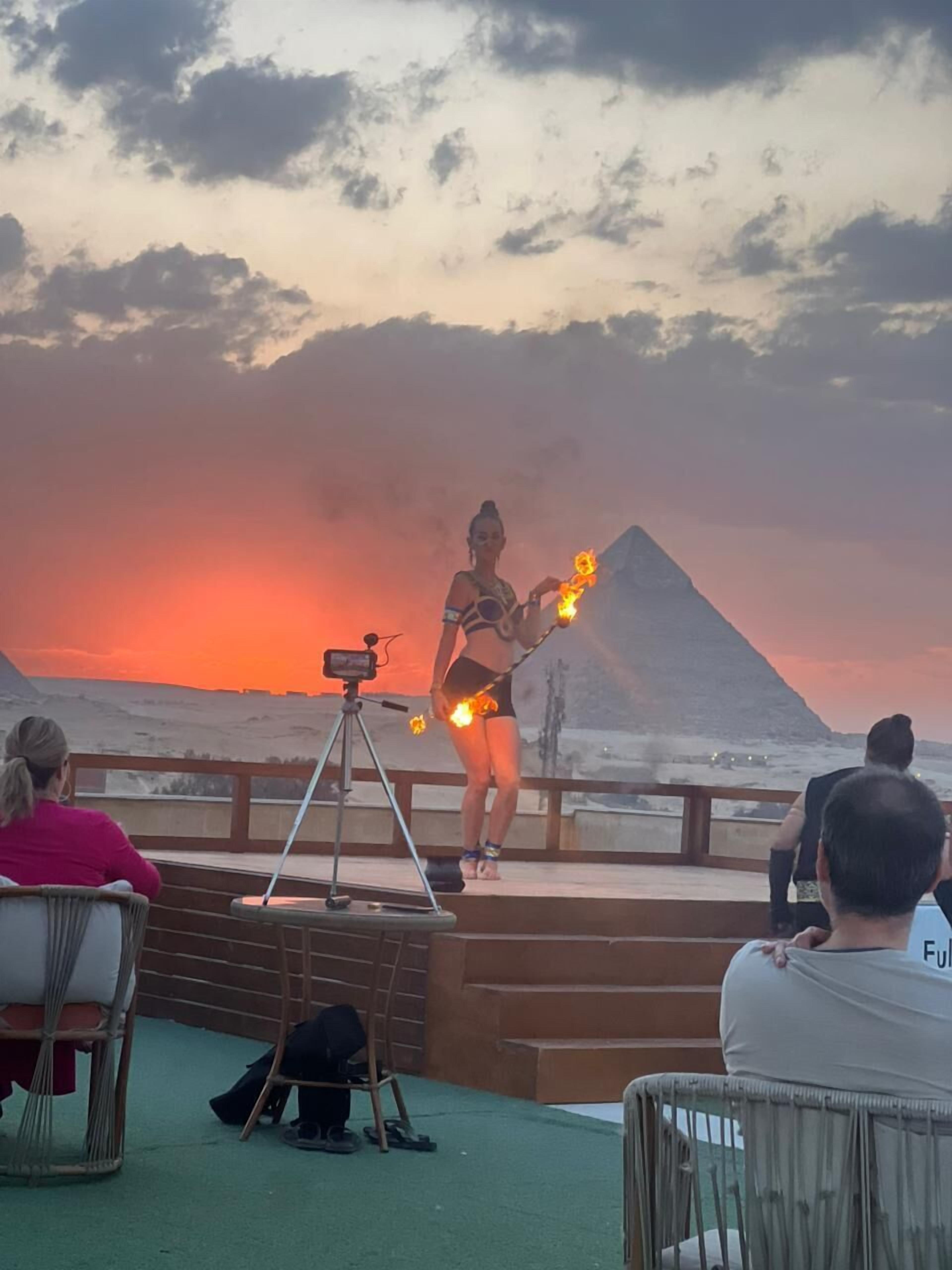 A person performing with fire on a stage with a backdrop of a volcano and sunset.