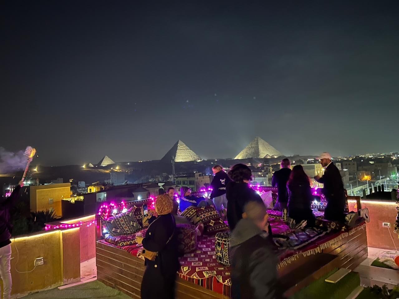 A rooftop terrace with people gathered around a table, overlooking a cityscape with pyramids in the background.