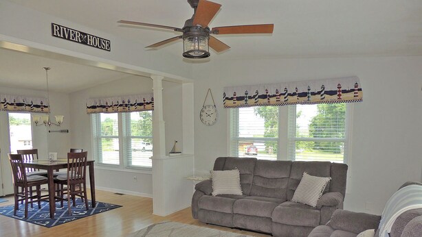 A living room with a gray sofa, a dining table with chairs, a ceiling fan, and a view of the outdoors through large windows. - Cute 3BR/2BA home located in the Village of Clayton with dock rental available (Clayton)