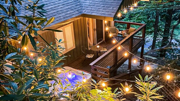 Une cabane en bois dotée d’une terrasse, entourée de verdure et illuminée de guirlandes lumineuses.