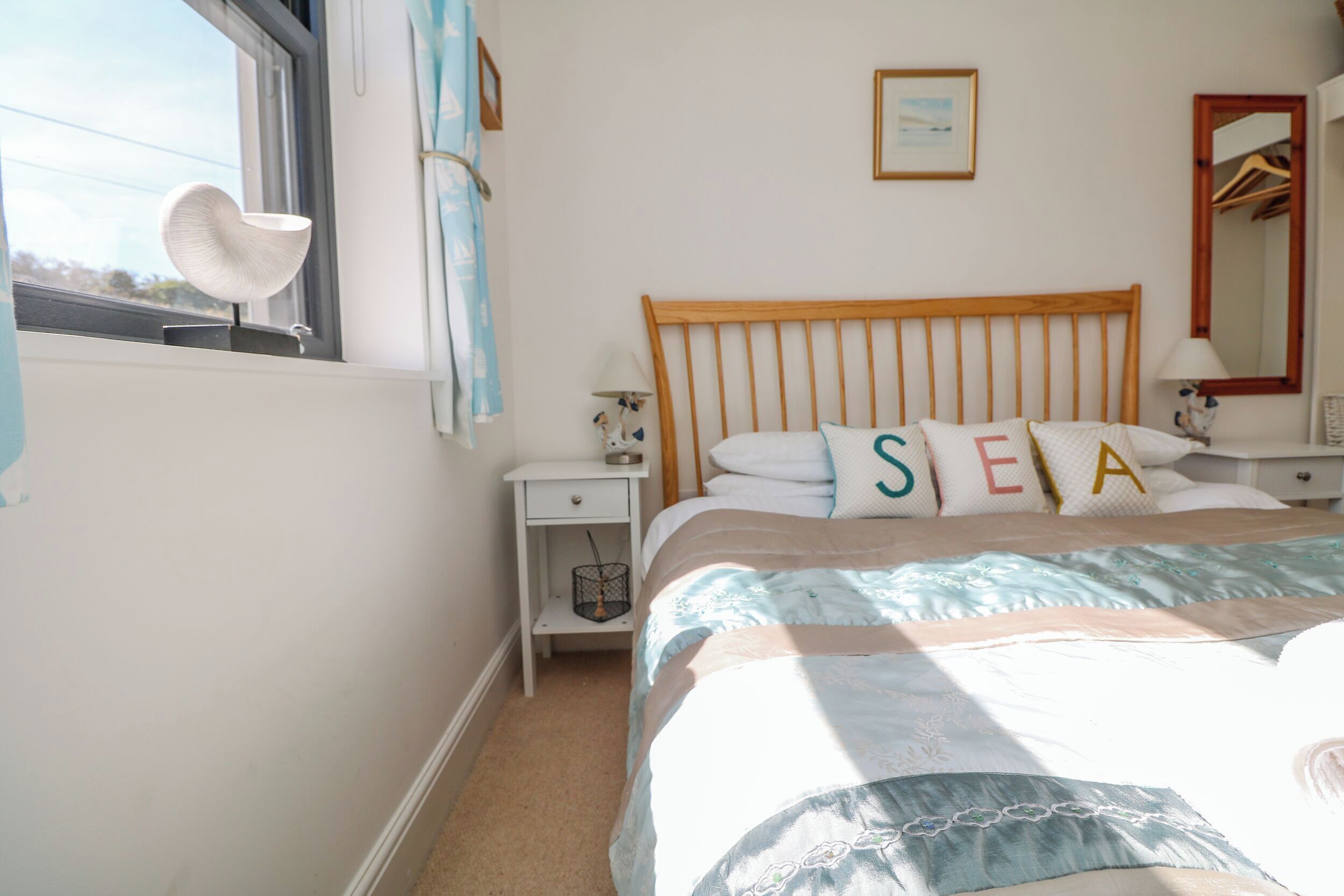 A bedroom with a wooden headboard, a bed with pillows spelling "SEA," a bedside table with a lamp, and a window with a decorative item.