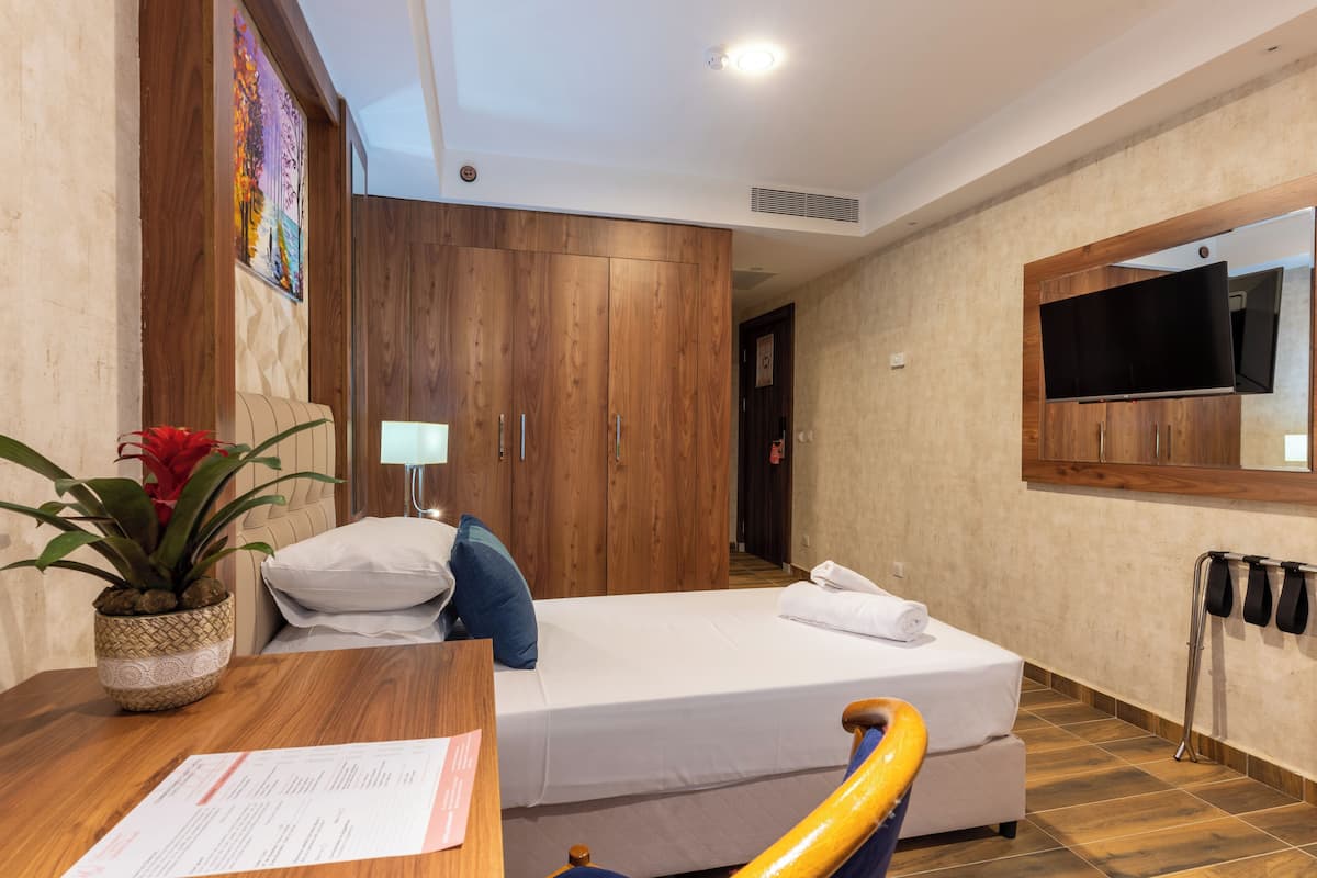 A hotel room with a bed, wooden wardrobe, a TV mounted on the wall, a potted plant, and a desk with a lamp.