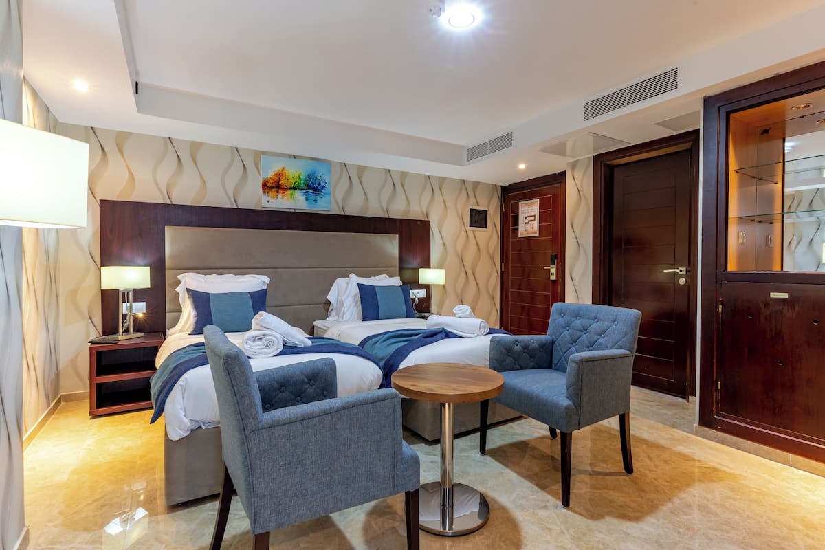 A hotel room with a large bed, two armchairs, a small round table, and a nightstand with a lamp.