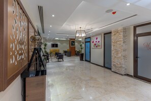 A spacious hotel lobby with a marble floor, a reception desk, seating area, and a wall-mounted TV.