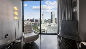 Ein modernes Hotelzimmer mit großem Fenster und Blick auf die Stadt, einem bequemen Sessel, einer Stehlampe und einem Flachbildfernseher.