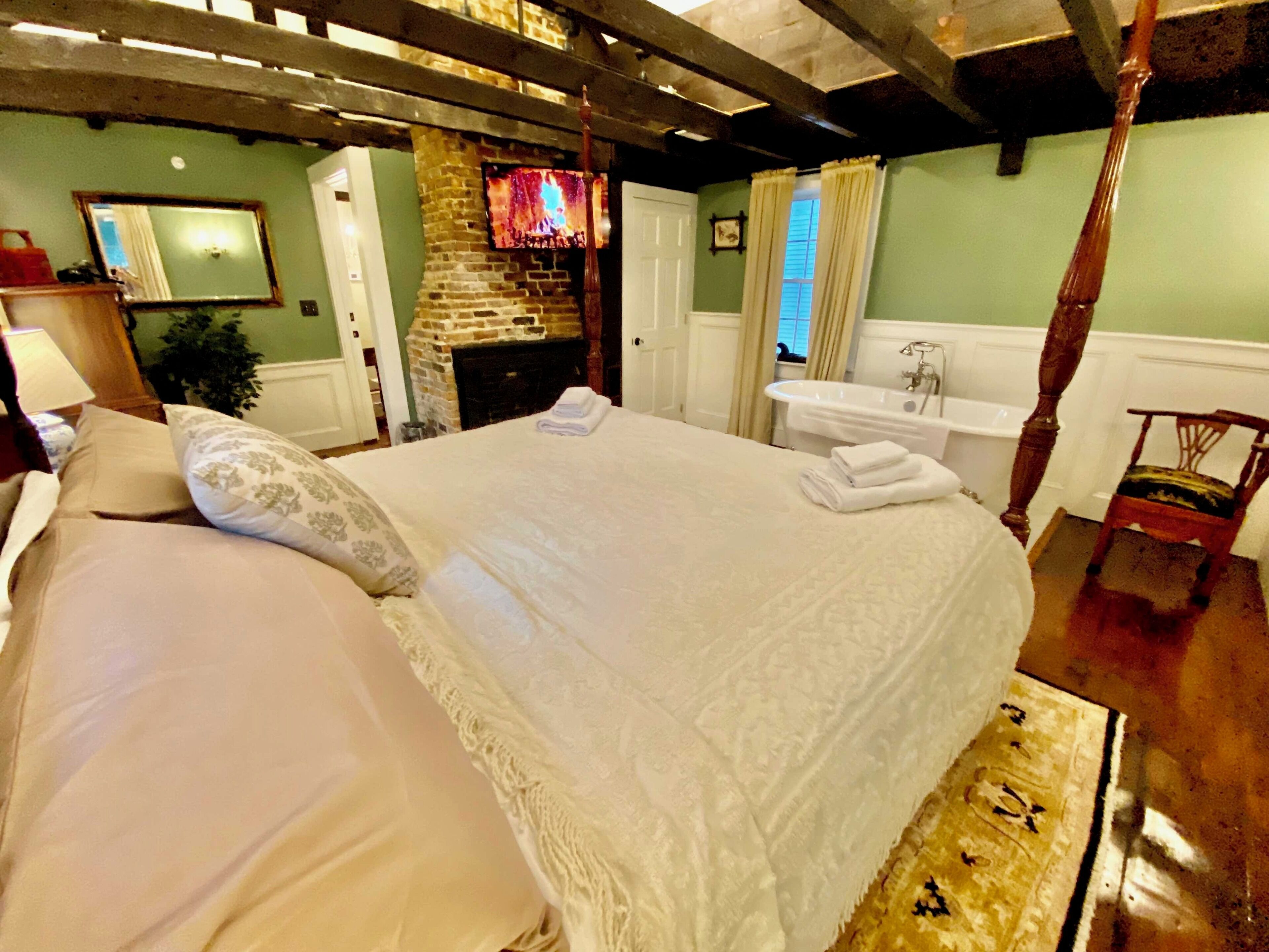 A bedroom with a large bed, a fireplace, a bathroom with a bathtub, and a wooden chair.