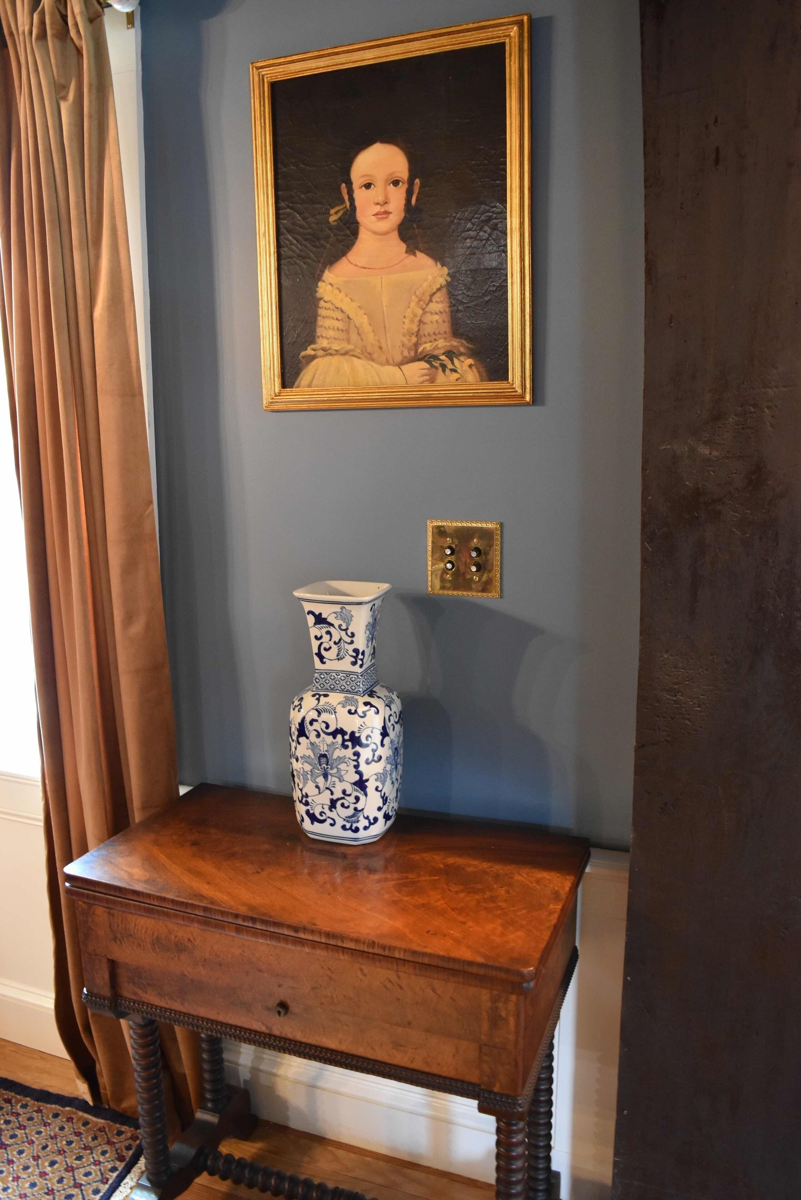 Een kamer met een blauwe wand, een lijst met een portret, een blauw-wit vaasje op een houten tafel, en een lichtschakelaar.
