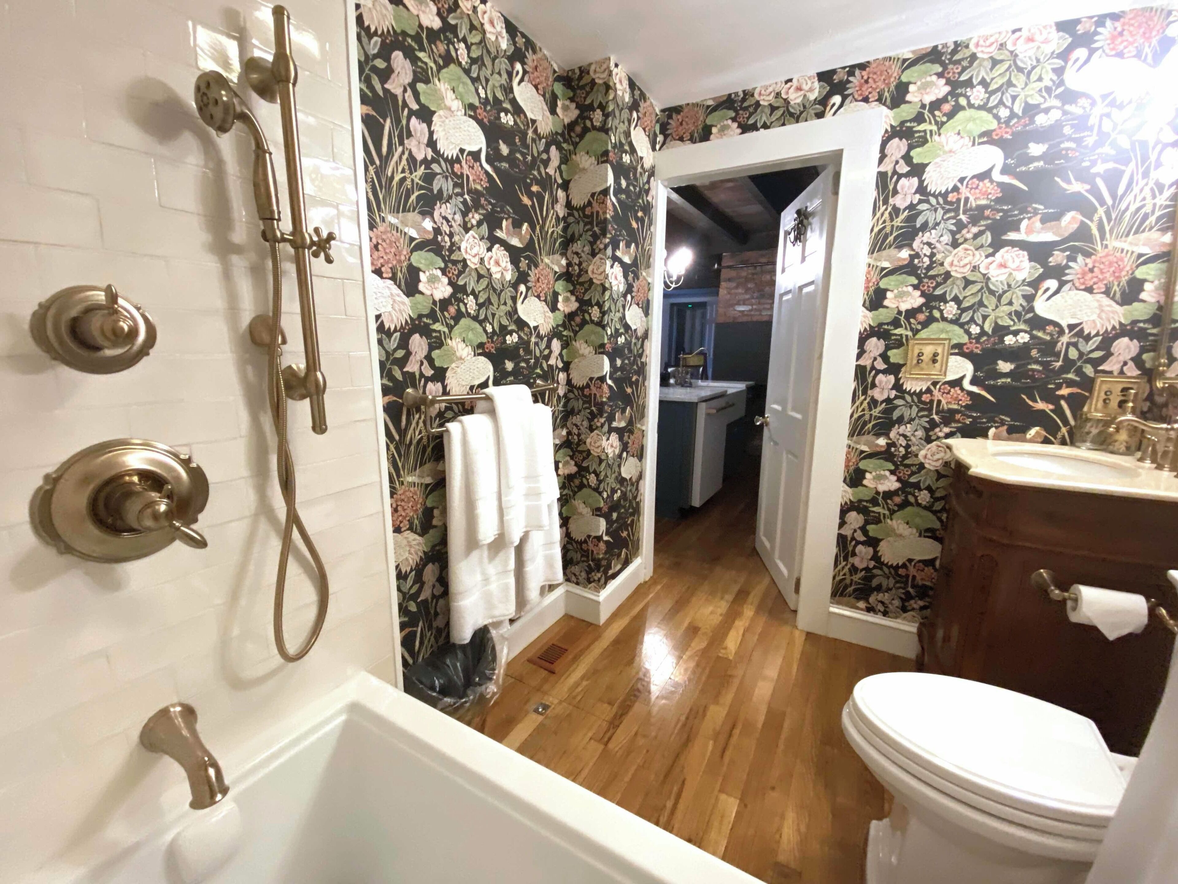 Un baño con ducha, inodoro y lavamanos. Presenta papel tapiz floral y suelo de madera.