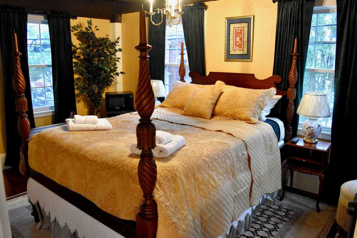 A four-poster bed with a gold and white bedspread, a large potted plant, and a framed picture on the wall.
