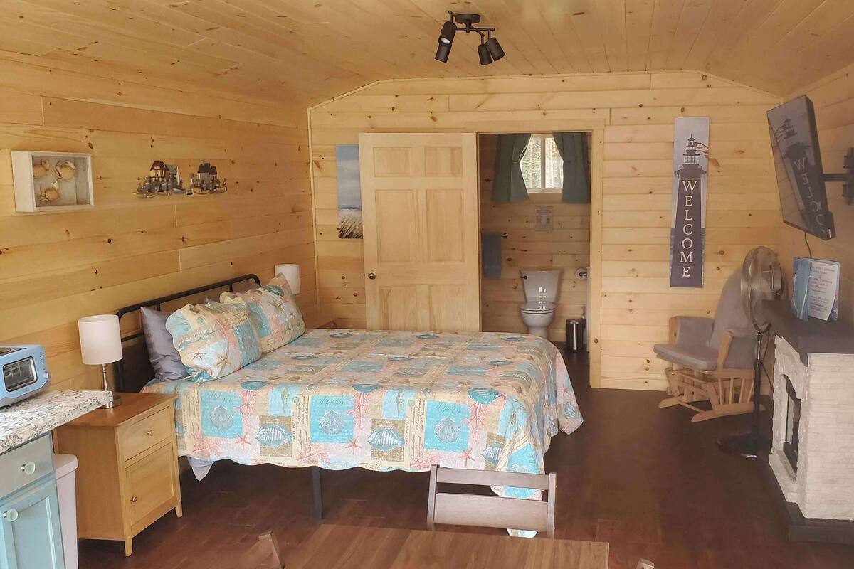 A cozy wooden cabin room with a bed, a small desk, a chair, a fan, and a sign that reads "WELCOME".