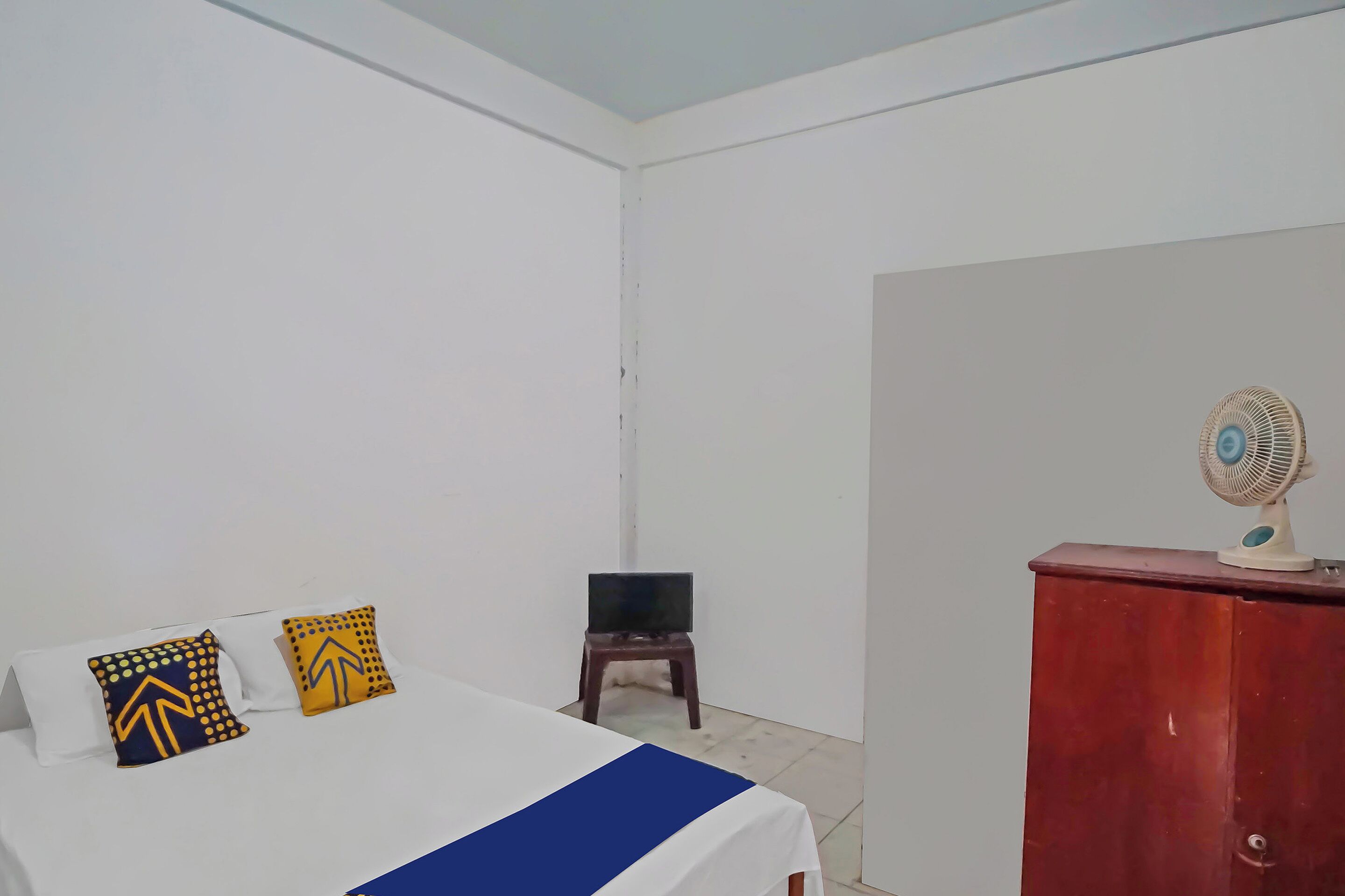 A bedroom with a bed, two pillows, a small table with a TV, and a fan on a cabinet.