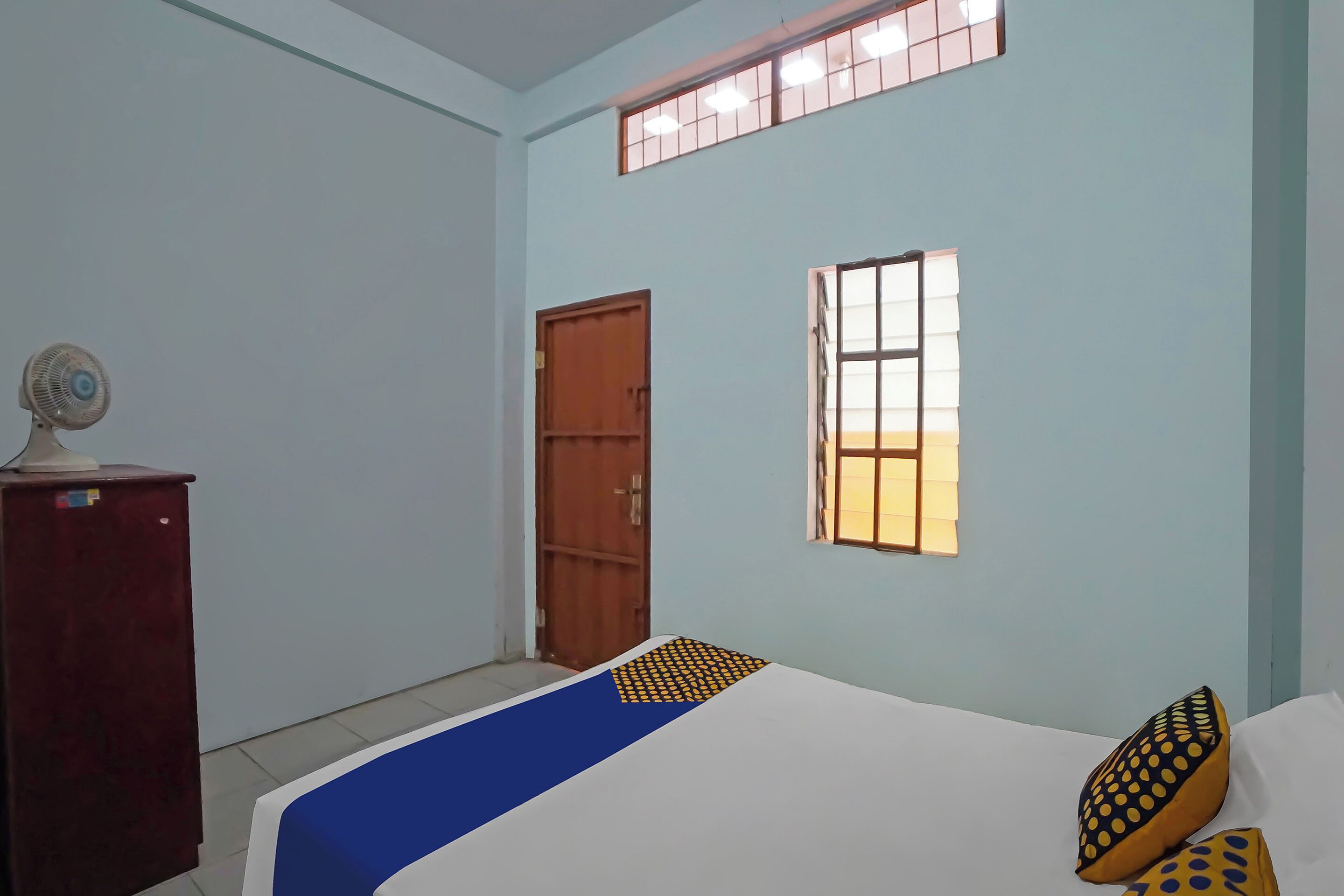 A room with a bed, a wooden door, a window, and a desk with a fan.