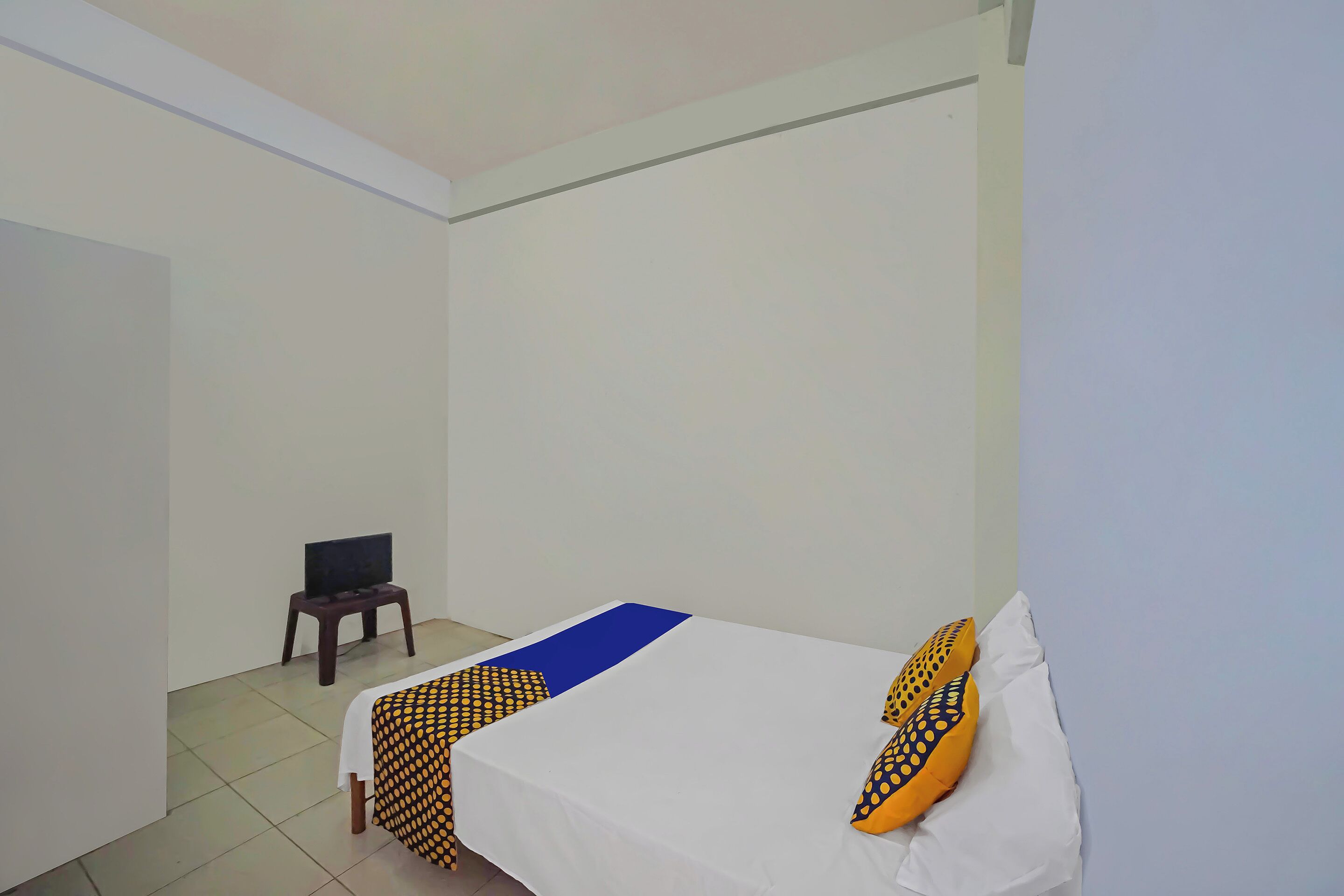 Una habitación pequeña y poco amueblada con una cama individual, un televisor sobre una mesita y una pared blanca sencilla.