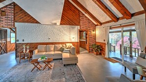 Un ampio soggiorno con un divano componibile, travi in legno e un'ampia porta scorrevole in vetro che si apre su un balcone con vista sugli alberi.