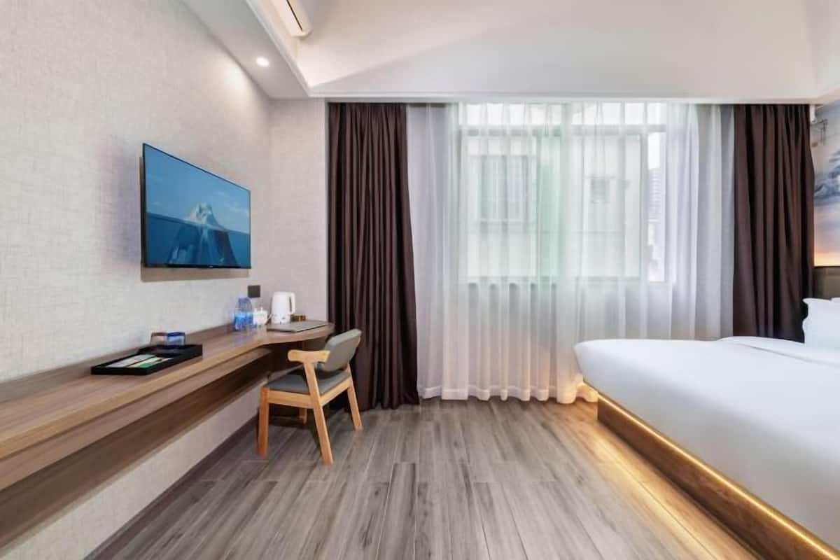 A hotel room with a bed, a desk with a chair, a flat-screen TV mounted on the wall, and a window with curtains.