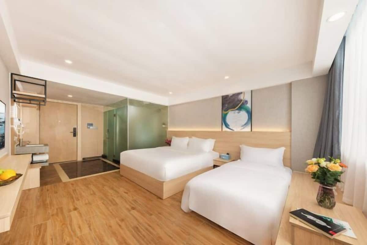 A hotel room with two beds, a desk, a chair, a small table, a vase with flowers, and a painting on the wall.