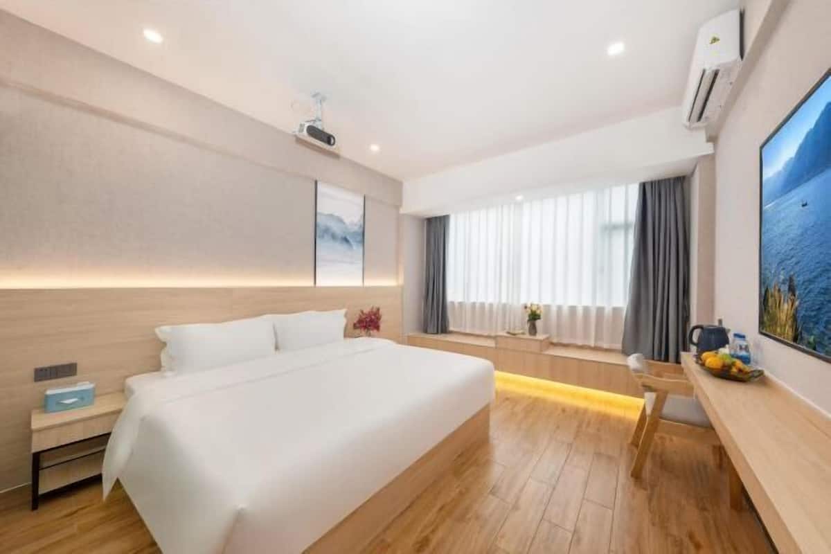 A modern hotel room with a large bed, a desk, a chair, a window with curtains, and a wall-mounted air conditioner.
