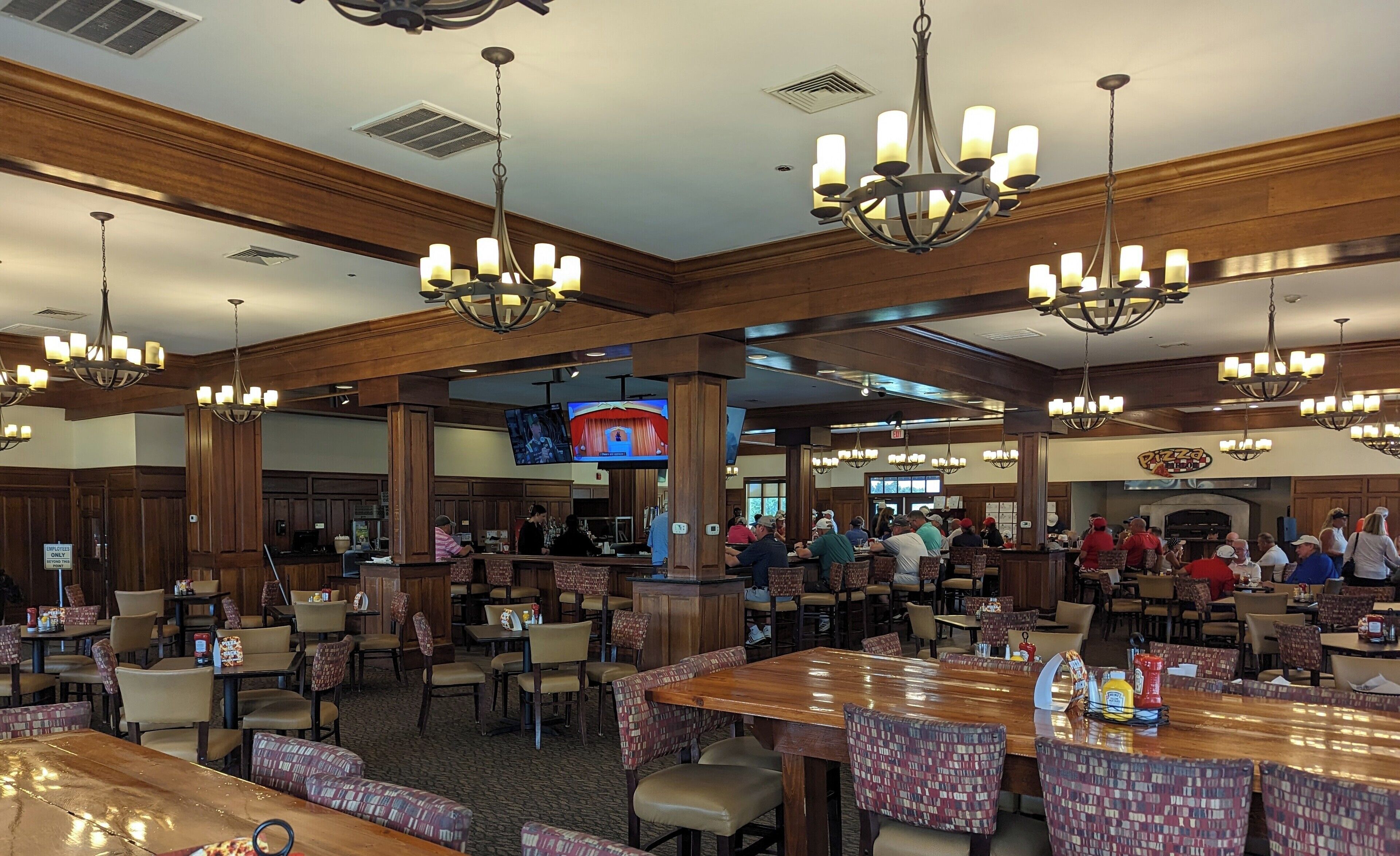 A spacious restaurant with high ceilings, wooden paneling, and multiple chandeliers. Tables are arranged in a casual layout, and a television is mounted on the wall.