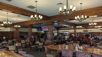 A spacious restaurant with high ceilings, wooden paneling, and multiple chandeliers. Tables are arranged in a casual layout, and a television is mounted on the wall.
