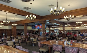 Un ristorante spazioso con soffitti alti, pannelli di legno e diversi lampadari. I tavoli sono disposti in modo informale e una televisione è montata a parete.