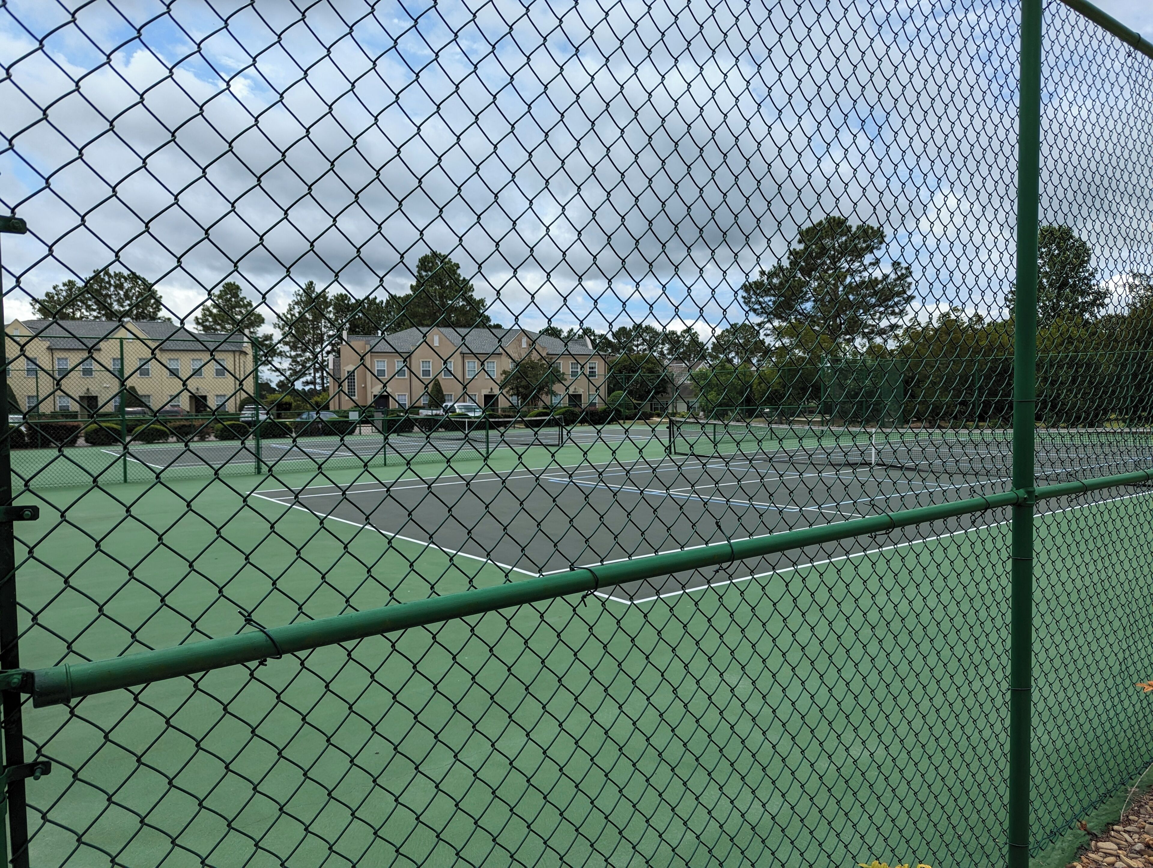 A chain-link fence surrounding a tennis court with a green surface, adjacent buildings, and trees in the background.