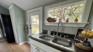 A kitchen with a double sink, a window with a view of trees, and a shelf with a plant and a mug. - Private Coastal Cottage on James Island (Charleston)