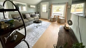 A living room with a sofa, coffee table, and a dining area with a table and chairs. There is a window with curtains, a ceiling light, and a mirror on the wall. - Private Coastal Cottage on James Island (Charleston)