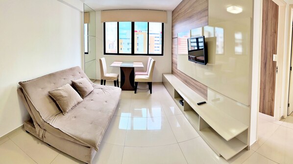 A modern living room with a sofa, a dining table, chairs, a TV, and a view of buildings through large windows.