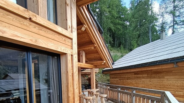 A wooden deck with outdoor seating, a glass door entrance, and a view of the surrounding forest.