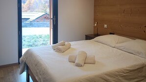 A bedroom with a wooden ceiling, a large bed with white linens, and a view of a forested area through an open door.