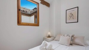 A hotel room with a bed, a nightstand with a lamp, a framed artwork on the wall, and a window with a view of a building. - Penthouse in Málaga by Costarentals - MAL01 (Málaga)