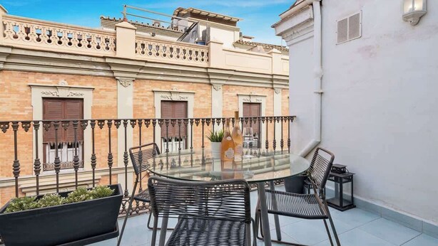 A glass-top table with four chairs on a balcony, overlooking a building with a wrought iron railing and a white wall with a light fixture. - Penthouse in Málaga by Costarentals - MAL01 (Málaga)