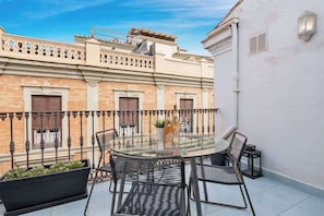 A glass-top table with four chairs on a balcony, overlooking a building with a wrought iron railing and a white wall with a light fixture.