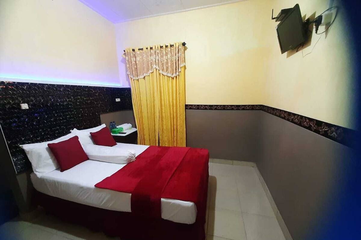 Una habitación de hotel con una cama, dos almohadas rojas, una colcha blanca, una manta roja, una cortina amarilla, una mesita con un objeto verde y un televisor instalado en la pared.