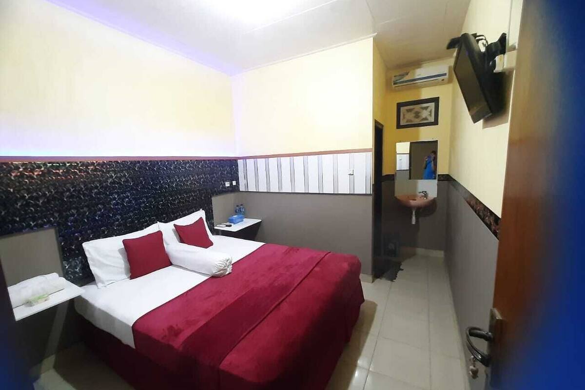 Habitación de hotel con cama, mesita de noche, televisor montado en la pared y baño visible a través de una puerta abierta.
