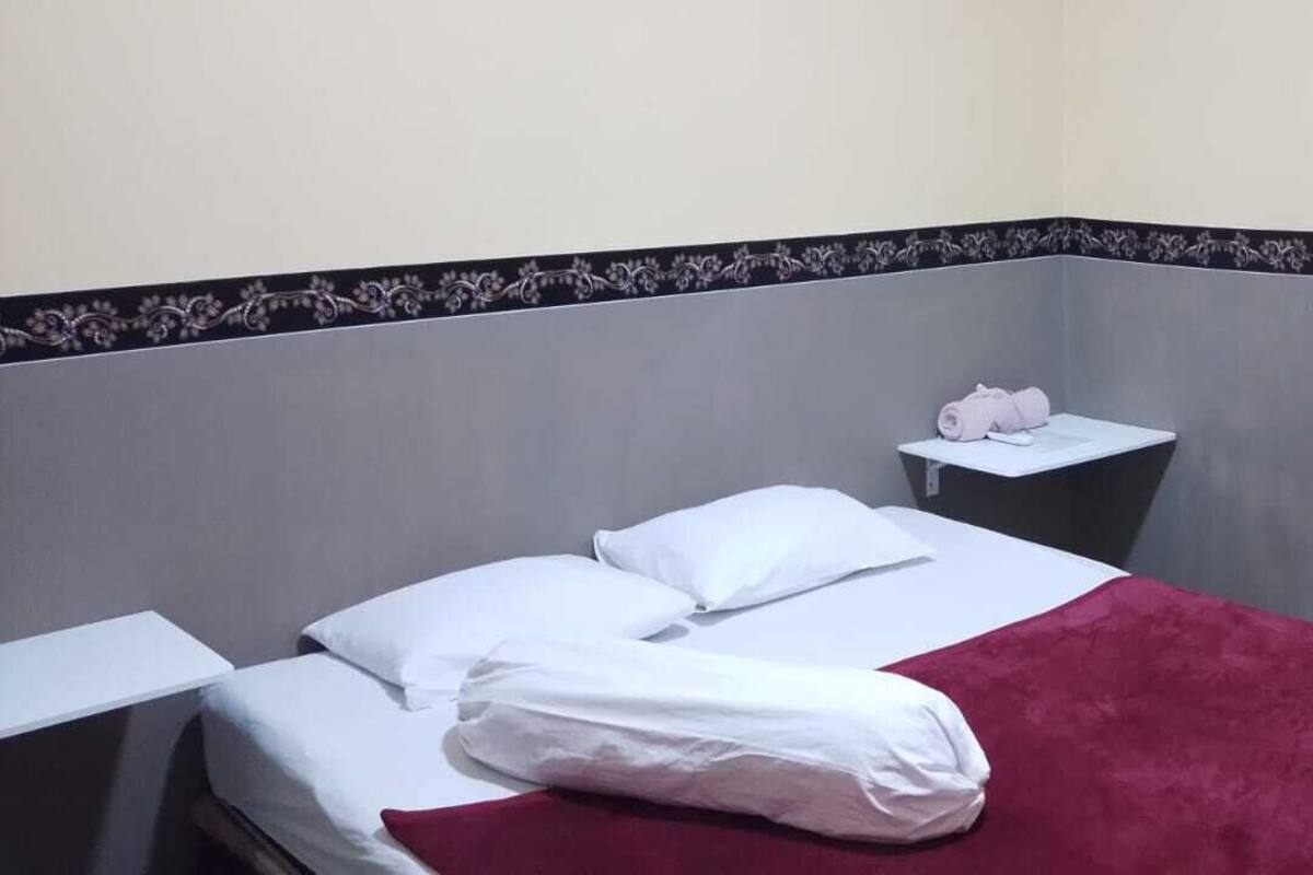 Una cama individual con sábanas blancas y una colcha roja, una mesita de noche con una toalla rosa, y una pared con un borde decorativo.