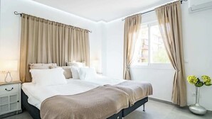 A modern bedroom with a large bed, beige curtains, a bedside table with a lamp, and a vase with flowers. - Beachclose apt Marbella by Costarentals - M28 (Marbella)