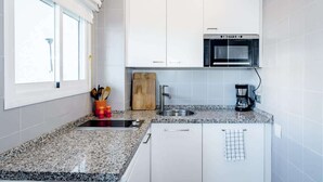 Une cuisine moderne avec des meubles blancs, un four encastré, une machine à café et une fenêtre équipée de stores.