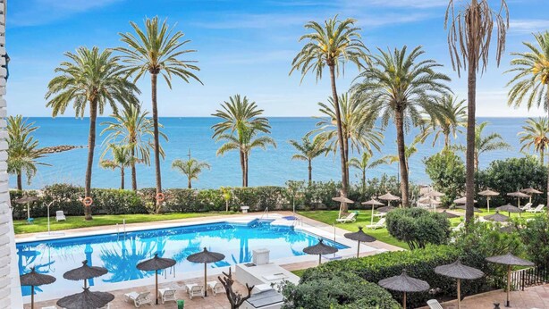Un espace piscine agrémenté de palmiers, un ciel d’un bleu limpide et une vue sur la mer.